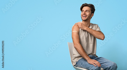 Immunization Campaign. Vaccinated Young Man Showing Arm With Adhesive Bandage After Vaccine Injection And Looking Away, Smiling Guy Sitting On Chair Over Blue Studio Background, Panorama, Copy Space
