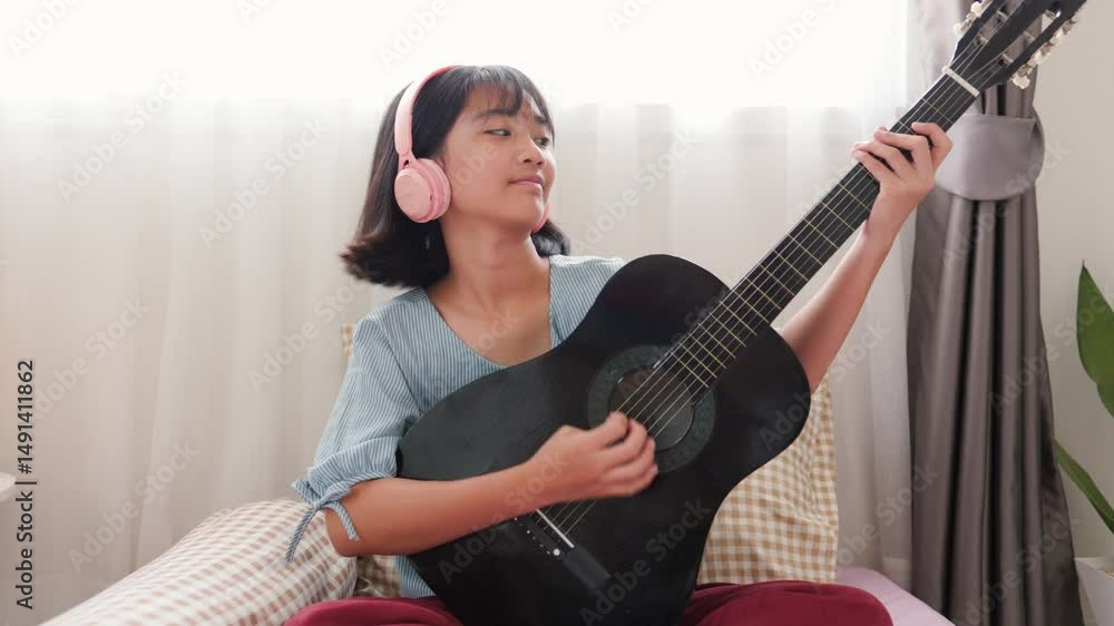Asian girl wearing headphones learning to play guitar at home using smartphone, showing modern self-learning and music practice in a cozy bedroom. Asian girl wearing headphones learning to play guitar at home using smartphone, showing modern self-learning and music practice in a cozy bedroom.