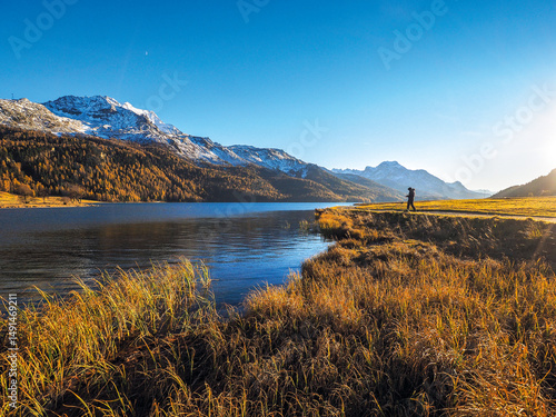 Lone hiker by calm alpine lake with autumn grass and snowy mountains under golden evening sunlight.  Beautiful landscape in the Swiss Alps on a day in autumn.