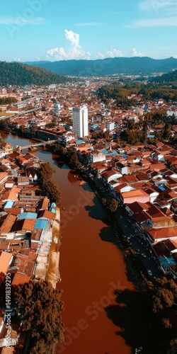 Aerial view of a city with a river running through it, surrounded by houses, buildings, and distant hills under a blue sky.