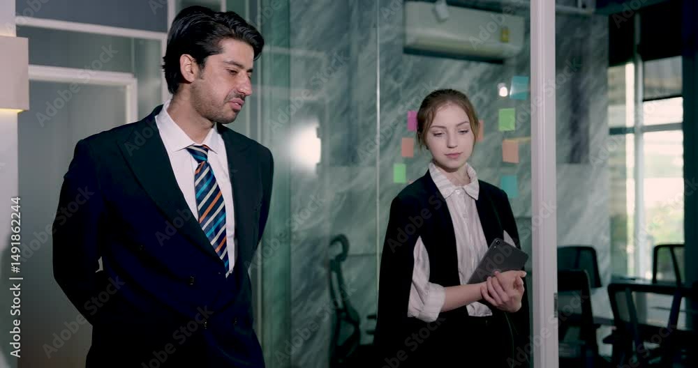 Businessman and businesswoman walking in office hallway talking about work plan and strategy while holding tablet expressing teamwork collaboration communication and business coordination success Businessman and businesswoman walking in office hallway talking about work plan and strategy while holding tablet expressing teamwork collaboration communication and business coordination success