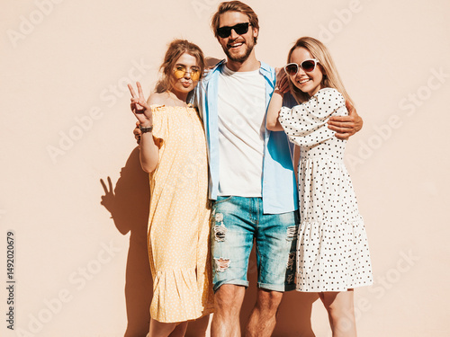 Group of young three stylish friends posing in the street. Fashion man and two cute female dressed in casual summer clothes. Smiling models having fun in sunglasses.Cheerful women and guy outdoors