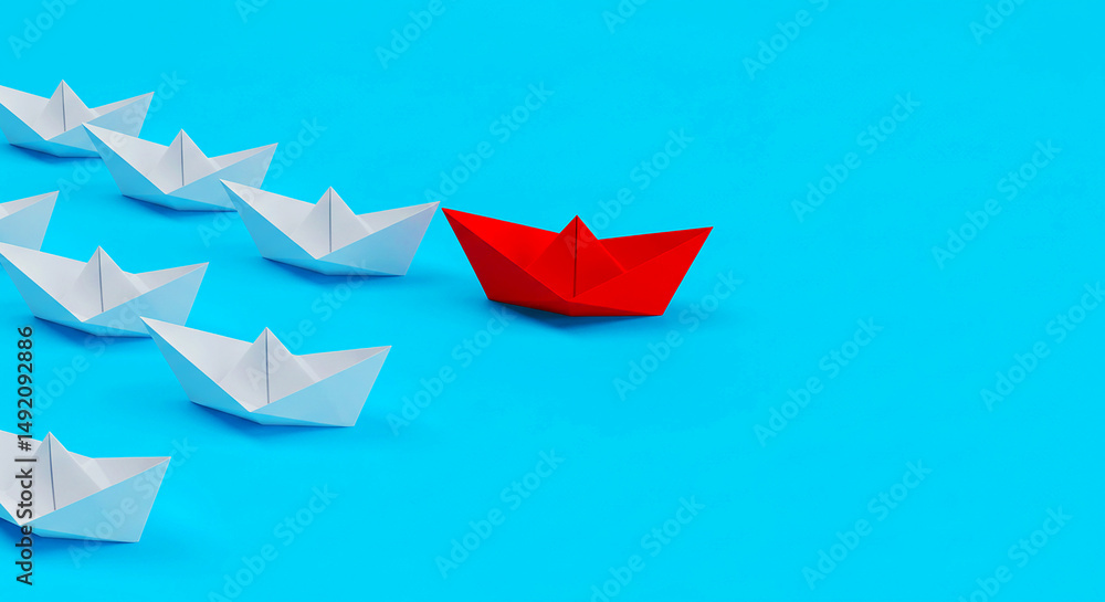 A red paper boat leading a group of white paper boats on a bright blue background in a studio shot A red paper boat leading a group of white paper boats on a bright blue background in a studio shot