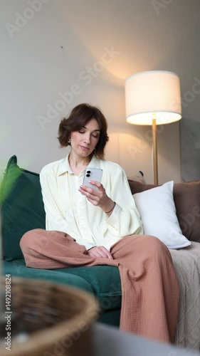 happy girl woman sitting at home on the couch looking at her phone and scrolling through her news feed. cozy living room interior