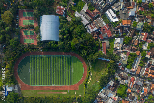 High angle aerial shot of a sports complex featuring a football field, tennis courts, and a large building, surrounded by trees and dense urban housing.