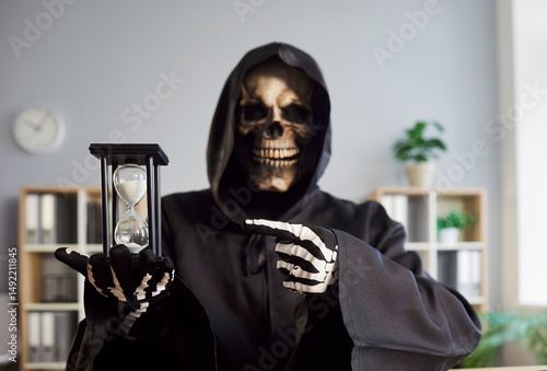 Photography Grim reaper of death in dark hooded cloak holding hourglass in skeletal hand and pointing at time