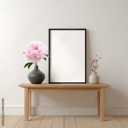 A stylish black frame stands on a wooden table, showcasing an empty white poster. Next to it are two decorative vases with a peony and delicate branches, adding charm to the cozy room