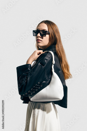 A stylish woman in a leather jacket with a bag in her hand poses against a light background