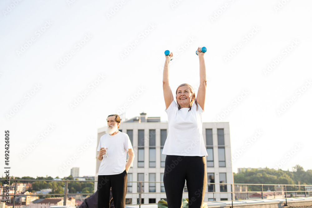 Fototapeta premium Elderly couple exercising together outdoors in urban area, woman lifting dumbbells, man observing. Healthy lifestyle, fitness, staying active in middle age.