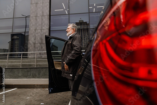 A middle-aged Caucasian man with a gray beard stands outside a parked car in an urban area, wearing a black jacket and exiting the vehicle in front of a modern glass building.