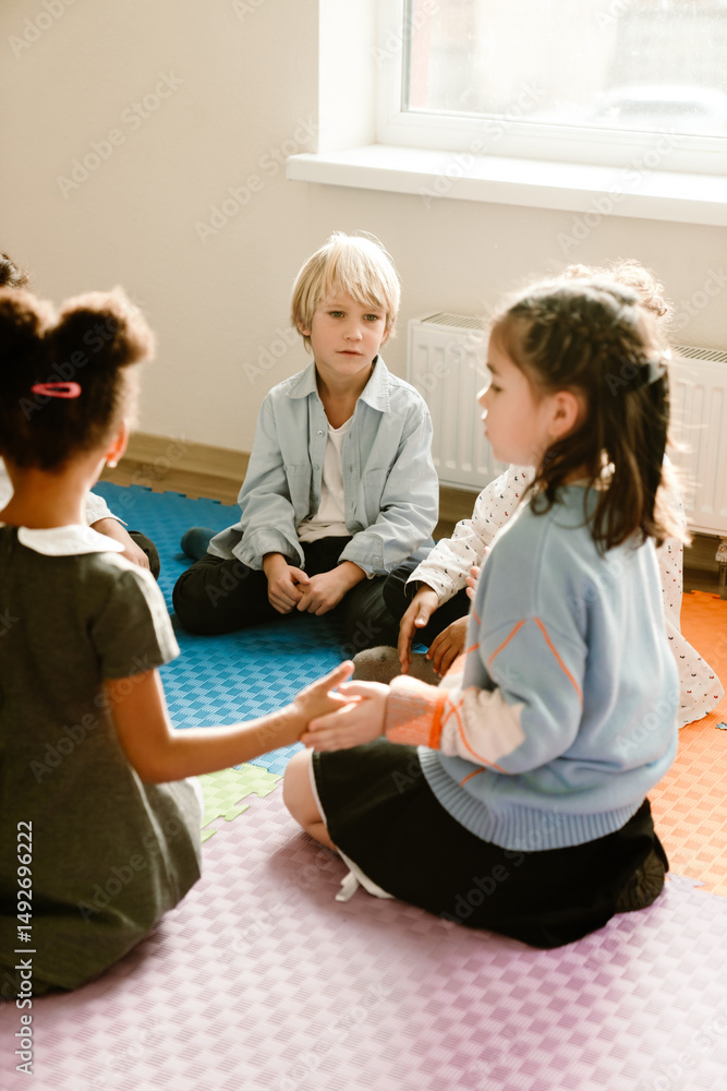 Obraz premium A school-aged White boy looks at his White girl classmate as they sit together with others in a circle on mats playing a game. A concept of childhood, communication, and the learning process.