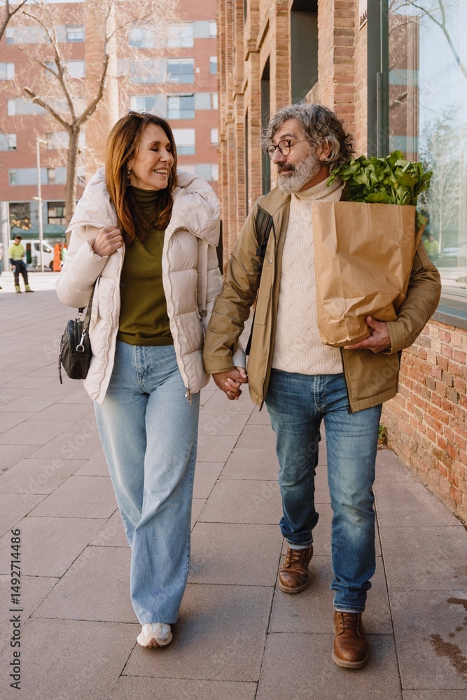Naklejka premium A White middle-aged man in eyeglasses with a beard is holding a paper shopping bag with parsley, and walking on a street with high buildings while holding his White middle-aged wife's hand
