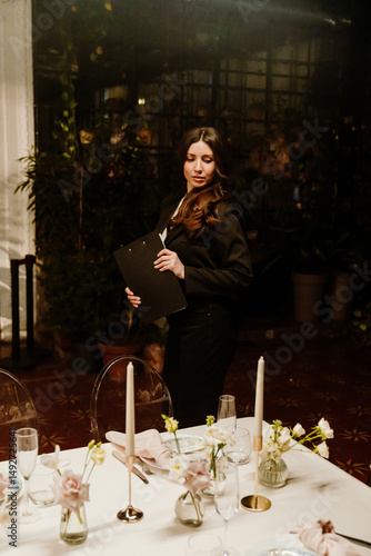 Принти на полотні A young White female wedding planner is standing beside a candlelit table with elegant white linens, floral arrangements, and champagne glasses while holding a clipboard in a dim indoor venue