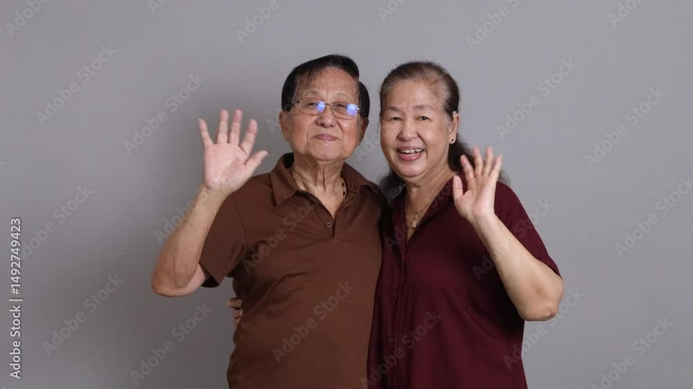 A senior East Asian couple in casual clothes standing against a grey background. A senior East Asian couple in casual clothes standing against a grey background.
