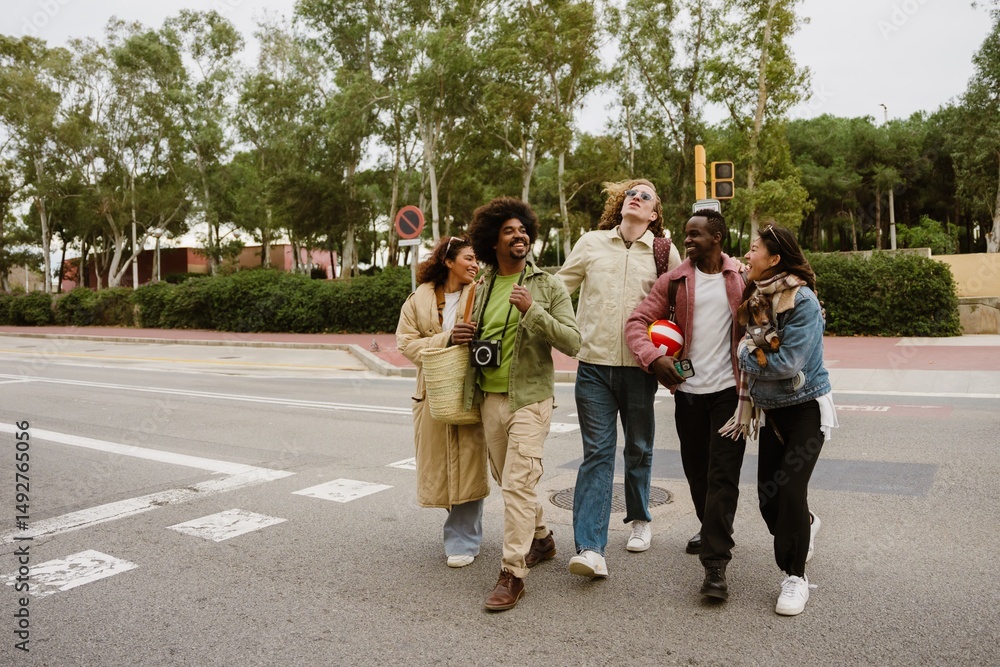 Obraz premium A multinational group of young people crossing a city street together in a cheerful mood, one woman holding a dog, others carrying a volleyball and camera, surrounded by greenery and an urban park.