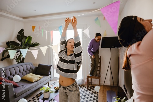 Three young, Asian and brown-skinned female friends decorate the living room for a party together, inflating balloons and hanging garland. A concept of teamwork, girlhood, and partying.