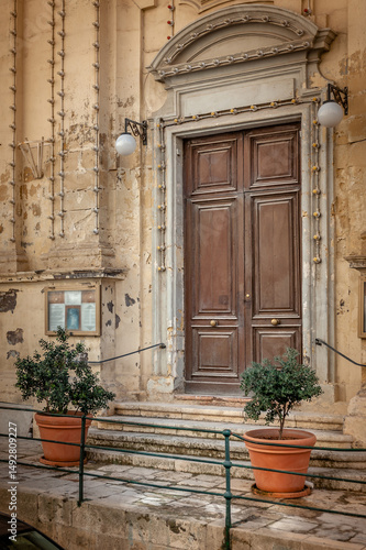 Traditional brown coloured wooden door. Close-up of the entrance to the church. The entrance is decorated with small potted trees. Mdina, Malta.