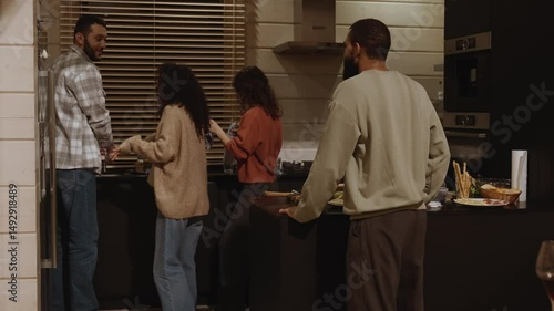Four adults, including a White man and a multiracial group, clean up together in a modern kitchen after a meal. They stand near the counter with dishes, food, and drinks.