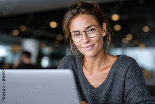A cheerful woman with glasses works on her laptop in a modern indoor setting  reflecting confidence  focus  and a blend of professionalism and approachability.