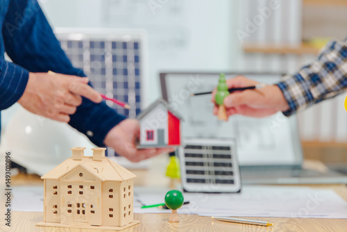 Architects and solar engineers discuss home design in office using model homes, solar panels, and architects' design documents. Concepts about home construction and solar power.