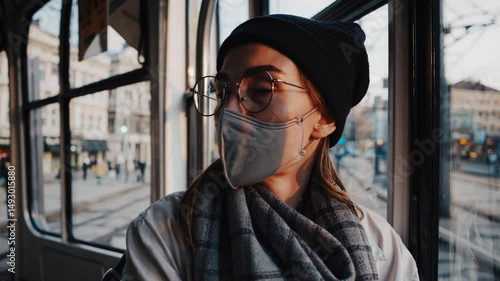 a young woman wearing a face mask looks out the window of a moving tram in a city setting.  The light casts a warm glow on her face.