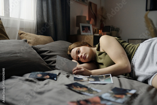 Konstfotografi Woman lying on bed in cozy room, holding and looking at photograph while surrounded by other photos