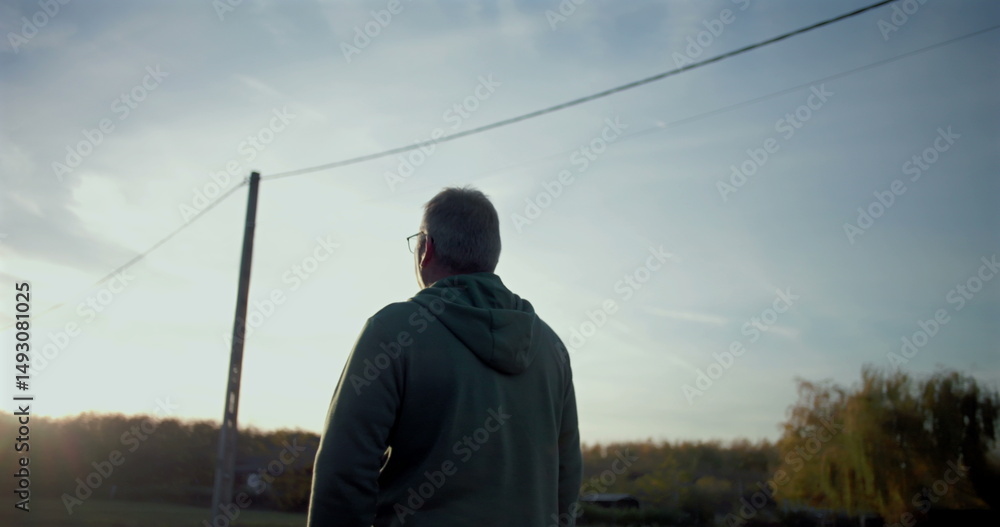 Fototapeta premium Man walking forward on a rural road, gazing ahead thoughtfully, serene open surroundings, utility poles and trees lining the path, quiet countryside atmosphere