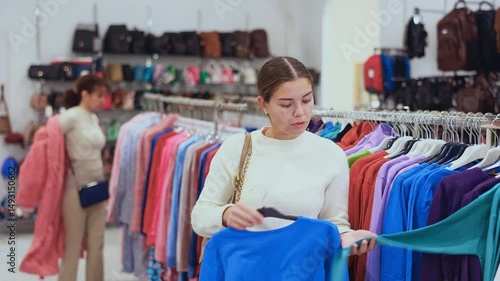 Girl buyer in store has removed hanger with long sleeve, from window and is considering product. Buyer mentally compares this piece of clothing and her wardrobe, fashion trends