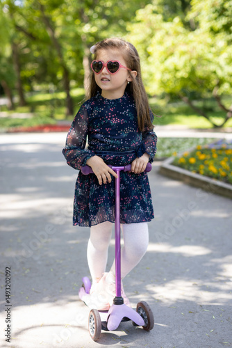 Smiling girl wearing heart-shaped sunglasses rides a scooter in a park. She enjoys a sunny day outdoors, representing childhood fun, happiness, and active lifestyle