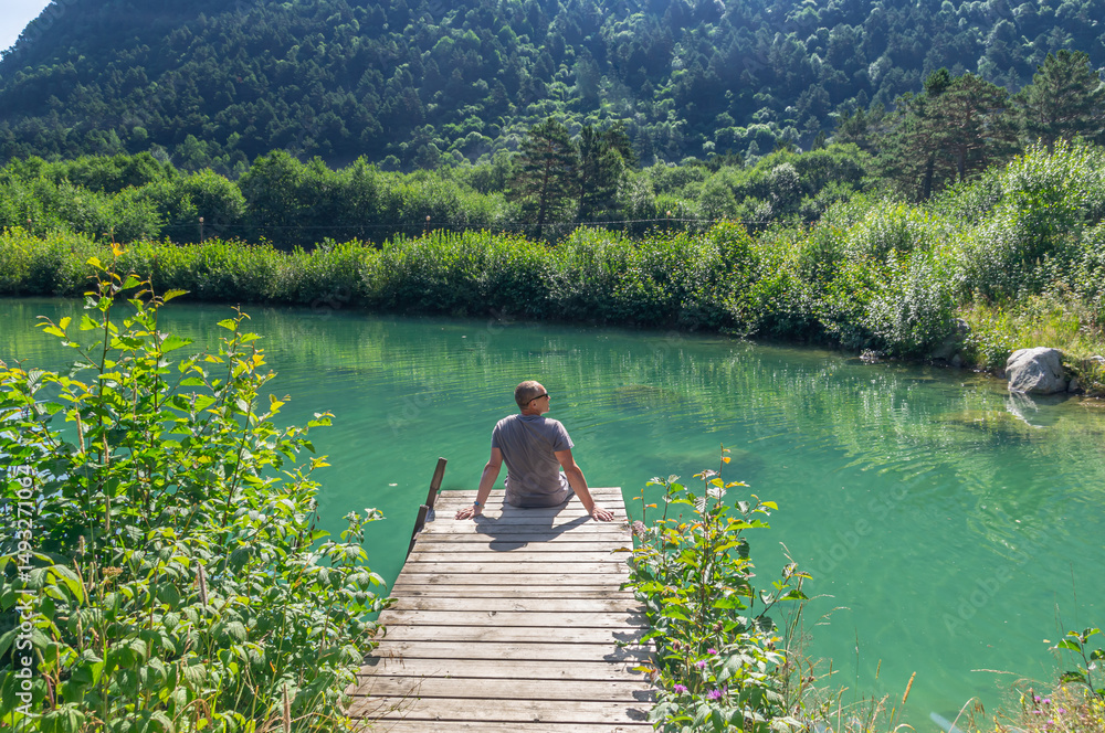 Fototapeta premium A man is sitting near a lake in the mountains. Summer vacation by the water. A man looks into the distance, sitting on the shore of the lake. The pond is overgrown with grass and shrubs.