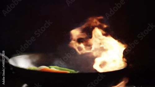Close-up of hand stir-frying vegetables in hot pan with flames, dynamic cooking scene with fresh ingredients and motion
