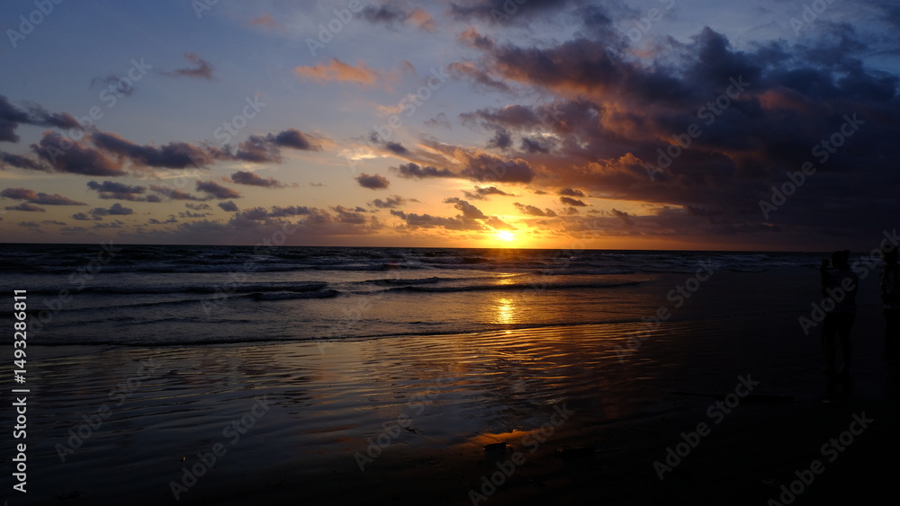 Naklejka premium A breathtaking sunset view at a beach in Bali, Indonesia, capturing the golden sun dipping into the ocean, dramatic clouds, and warm reflections on the wet shoreline.