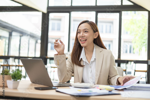A woman is working at a desk in an office with a laptop, a mobile phone and a pile of documents. She is smiling and happy.
