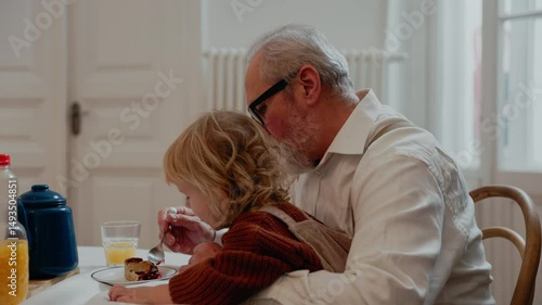 Grandson sits on grandfather's legs who is sitting at the table eating pie and feeding him