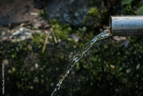 Billede på lærred Clear water streams from a metal pipe in a natural outdoor setting, with a soft-focus background of earthy tones and greenery
