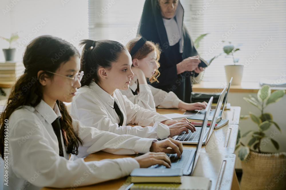 Obraz premium Students typing on laptops while being supervised by teacher. Classroom setting with natural elements like plants and light from windows providing calm environment