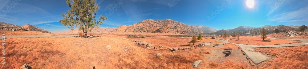 Fototapeta premium Panoramic view of a dry, sunlit mountain valley in California, featuring golden-red grass, winding paved paths, and scattered trees under a clear blue sky.