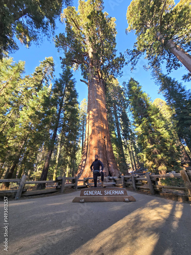 A man standing near the General Sherman Tree in Sequoia National Park, California. The image captures the scale and majesty of the world's largest tree alongside a human figure for perspective.