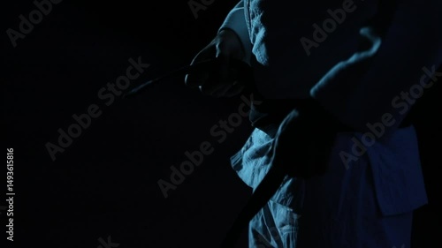 Close-up: Martial artist in gi meticulously ties their black belt in slow motion. Dark, moody atmosphere with cool blue lighting emphasizes focus and pre-fight ritual.