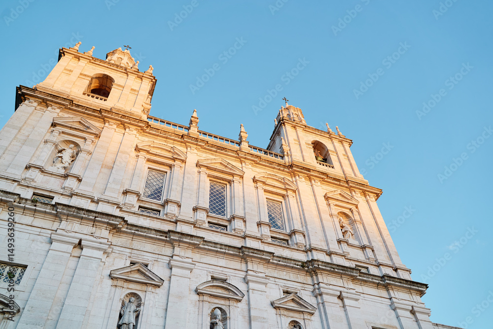 Obraz premium A close-up view of a historic church facade with intricate architectural details, featuring two tall towers and statues, illuminated by warm sunlight against a clear blue sky.