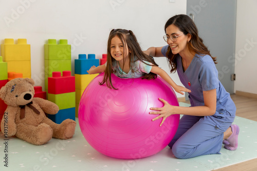 Pediatrician assisting girl balancing on exercise ball in clinic