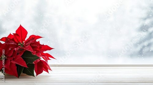 A vibrant red poinsettia sits on a rustic wooden table near a window looking out onto a snowy winter scene with falling flakes.