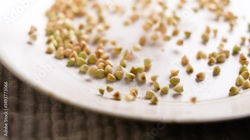 Green buckwheat falling on ceramic kitchen plate in slow motion close-up. Raw nutritious grain ingredient