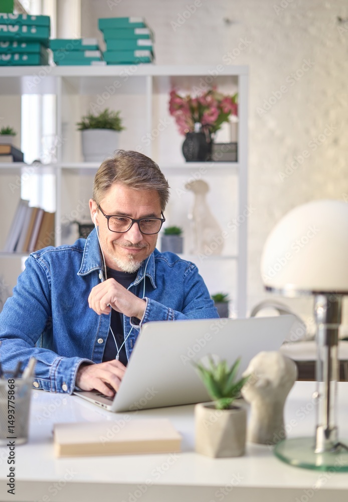 © nyul - Older man sitting at desk in bright room working with laptop computer in home office. Mature age, middle age, mid adult casual man in 50s, confident happy smiling.