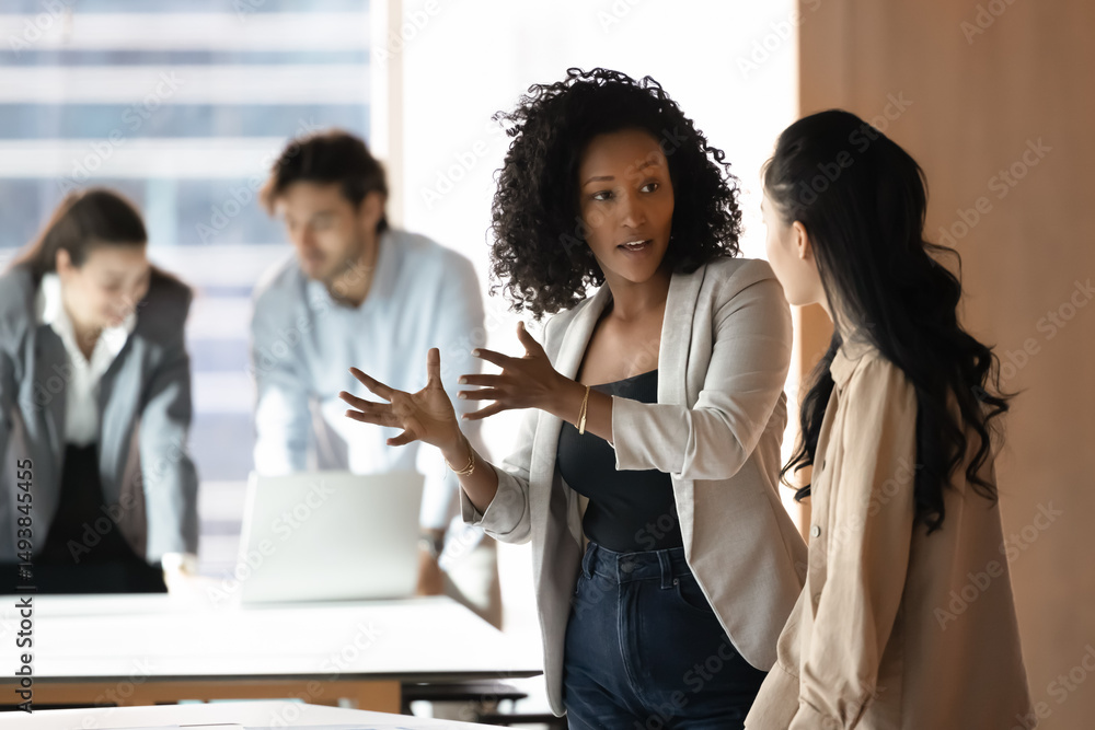 Obraz premium Two women engaged in focused conversation in office, African businesswoman speaking, explains idea or giving instructions to Asian female colleague during teamwork in workplace. Business communication