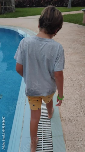 A boy is walking on a pool deck. He is wearing a grey shirt and yellow shorts. The pool is blue