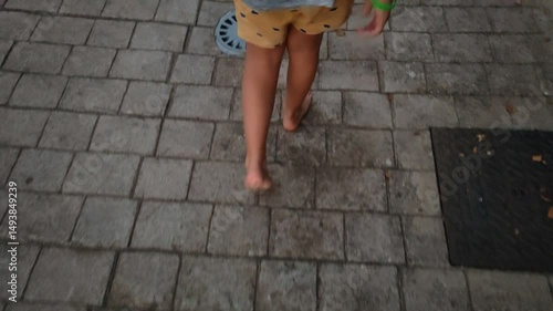 A child is walking barefoot on a brick sidewalk. The child is wearing shorts and a shirt