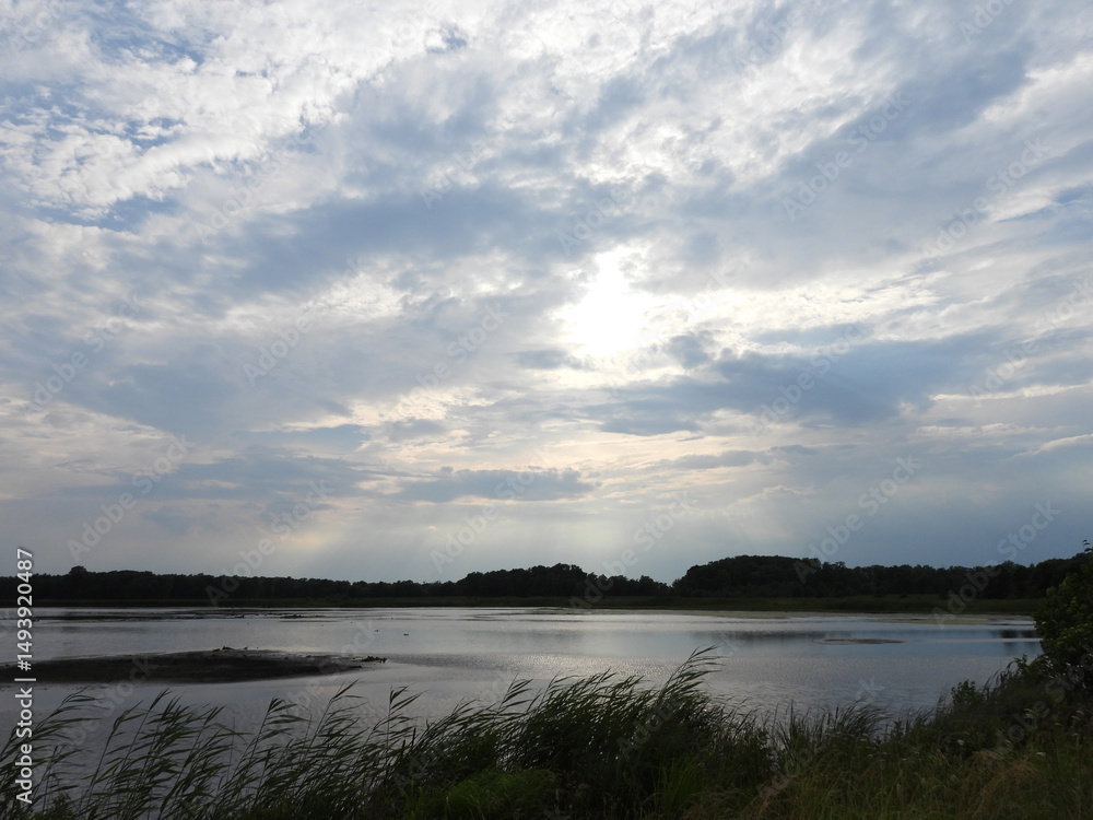Obraz premium Sunlight beams down from the clouds, highlighting the natural beauty of the wetlands within the Bombay Hook National Wildlife Refuge. Cloudy, summer day, Kent County, Delaware.