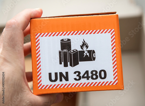 Paris, France - Apr 22, 2025: A closeup of a hand holding an orange box featuring a UN 3480 label with a warning image showing lithium batteries, one of which is on fire, indicating hazardous material