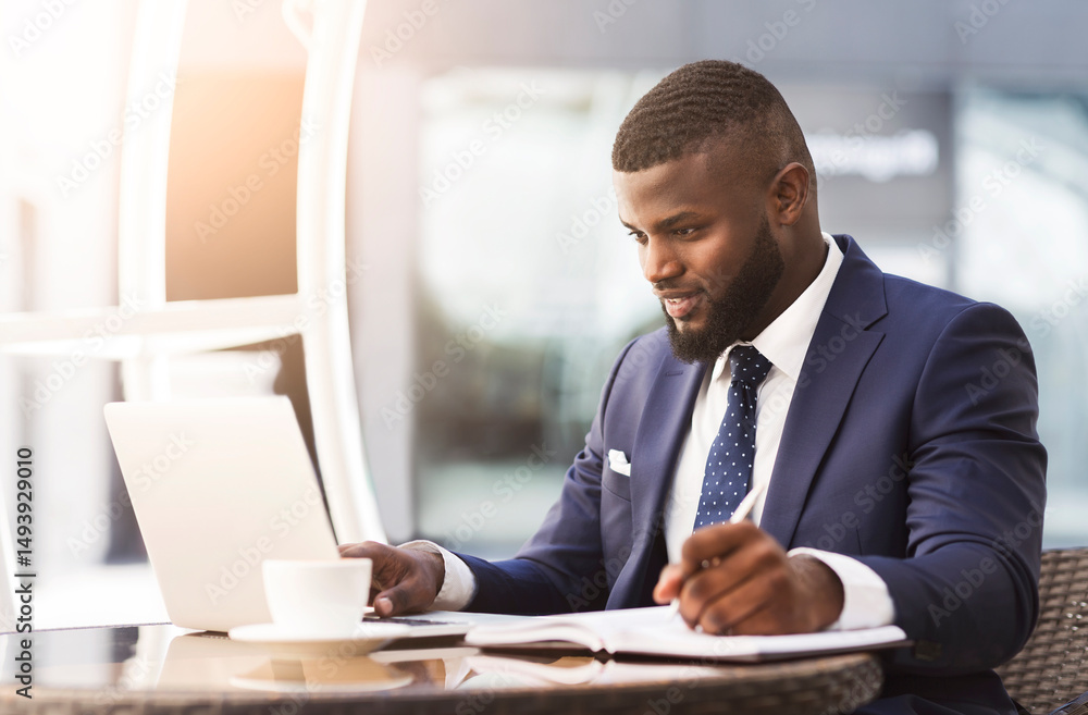 Fototapeta premium Serious Afro Businessman Working On Laptop Taking Notes During Coffee Break In Outdoor Cafe. Busy Day Concept
